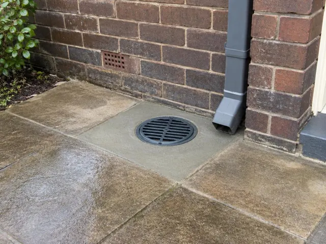 Outdoor gully grate set in wet paving beside a brick wall and downpipe in The Junction
