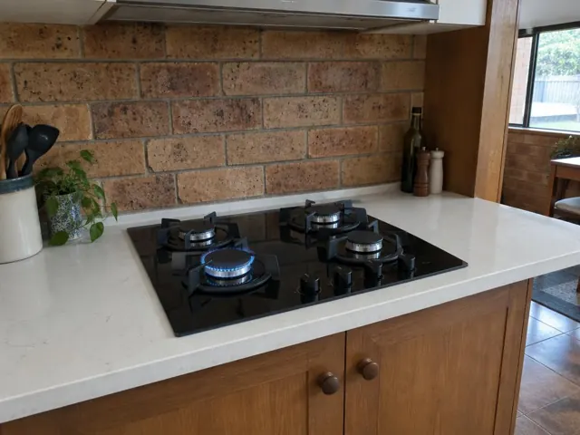Black gas cooktop set into a white benchtop with brick splashback in a timber kitchen in Whitebridge